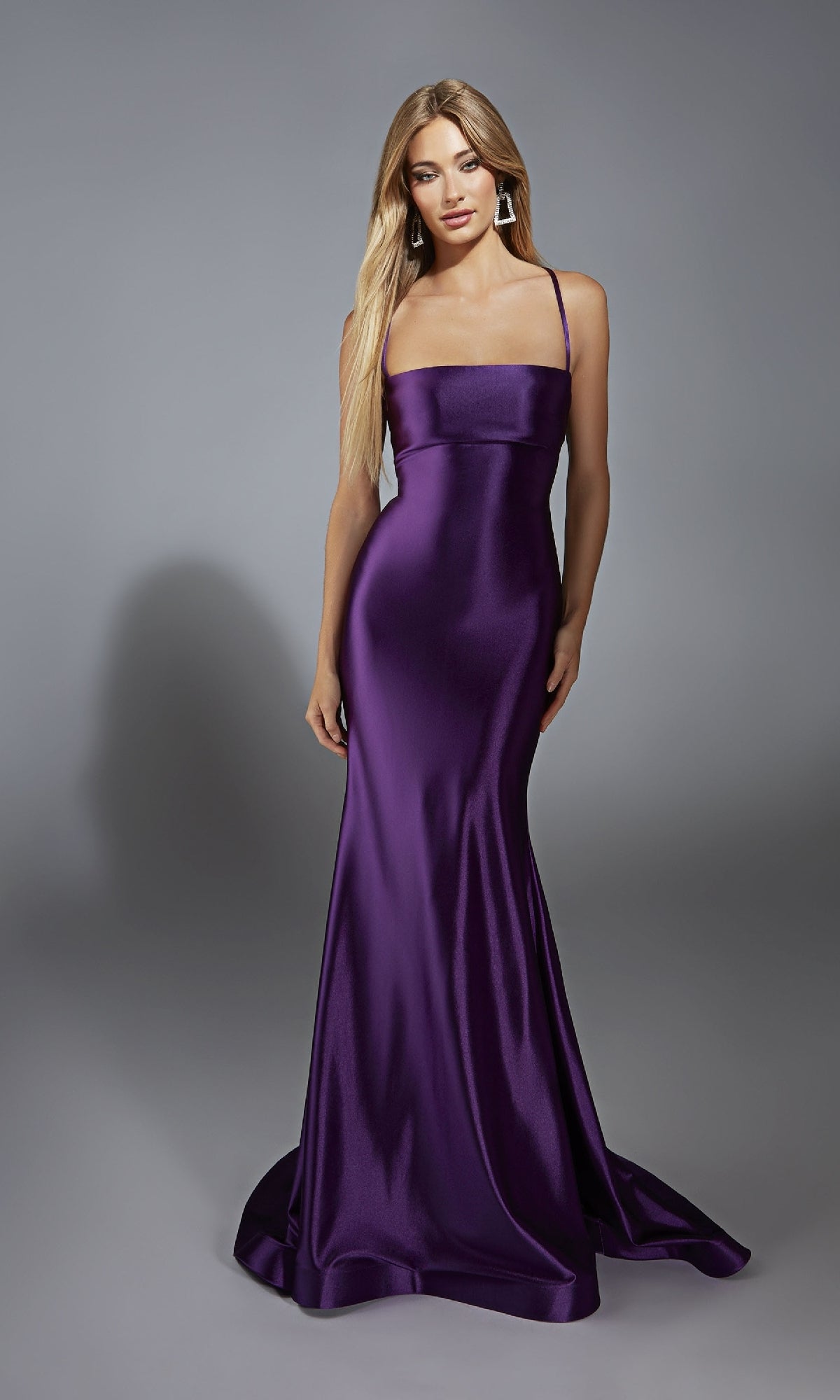 A woman poses against a gray background in the Aerie Couture 313, a floor-length, sleeveless satin prom dress in purple with lace-up open back, thin straps, and geometric earrings.